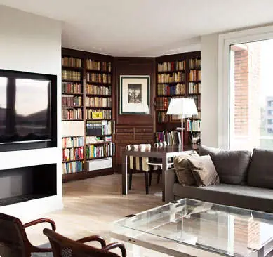 Salón con chimenea, biblioteca de madera oscura del suelo al techo, sofá gris y mesa de centro de vidrio.