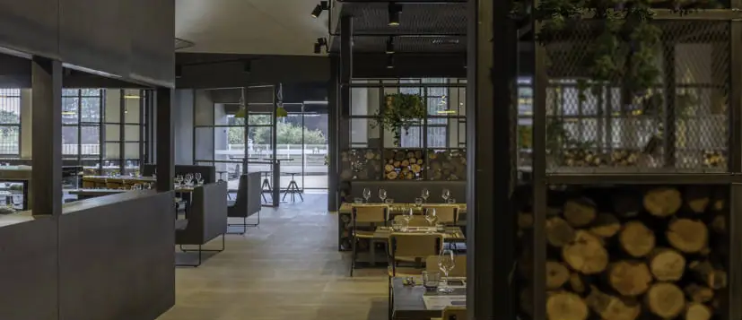 Comedor de La Industrial con mamparas de malla metálica, troncos de leña apilados y carpinterías metálicas