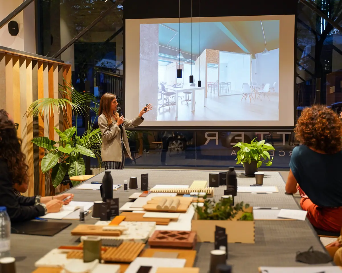 Una ponente presenta un proyecto de oficina proyectado en pantalla ante la mesa de materiales