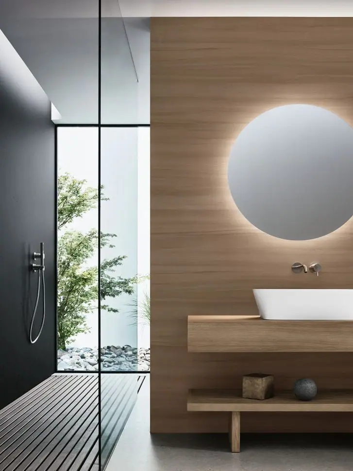 Baño con lavabo blanco sobre encimera de madera, espejo redondo retroiluminado y ducha abierta a un patio con arce y cantos rodados.