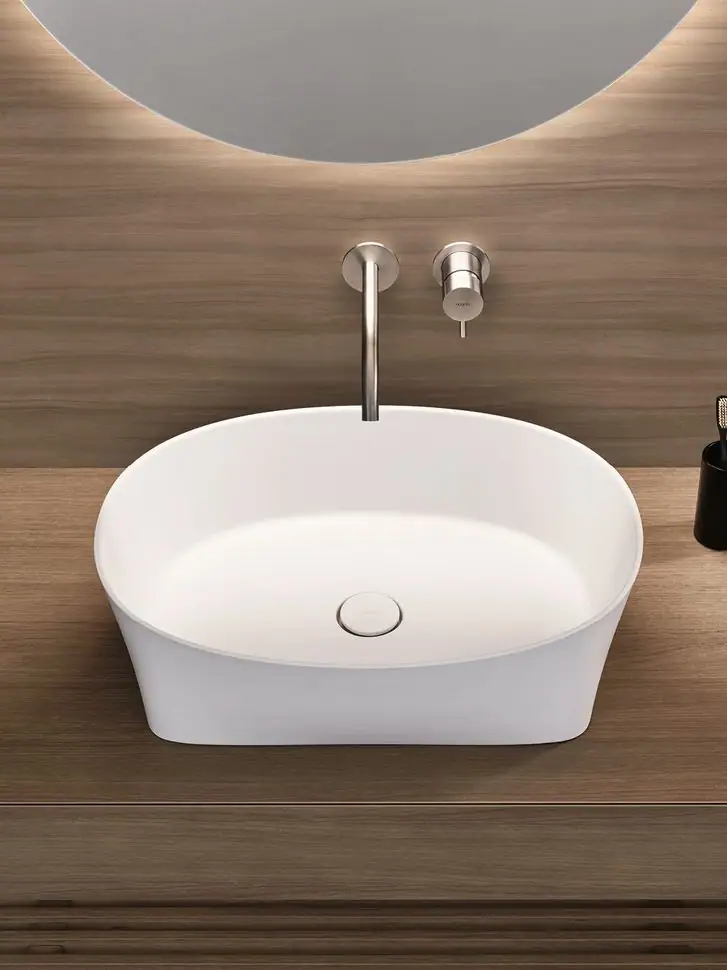 Vista superior de un lavabo blanco ovalado sobre encimera de madera, con grifería mural y espejo retroiluminado.