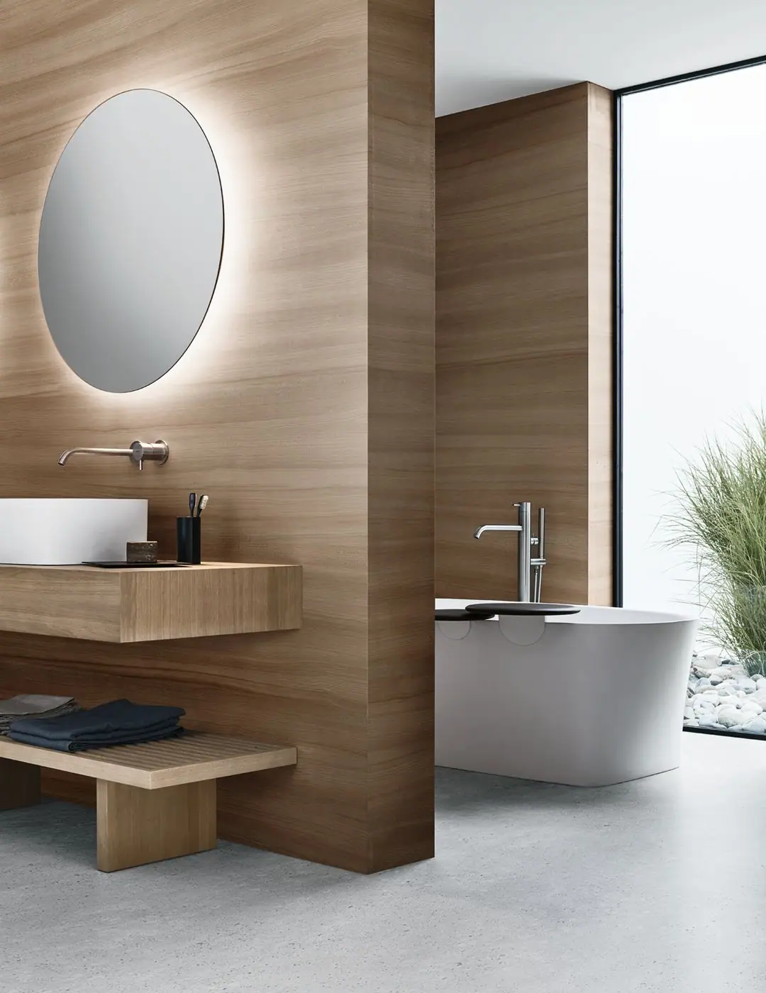 Baño con lavabo blanco sobre encimera de madera, espejo ovalado retroiluminado y, tras un tabique de madera, una bañera blanca exenta con bandejas oscuras.