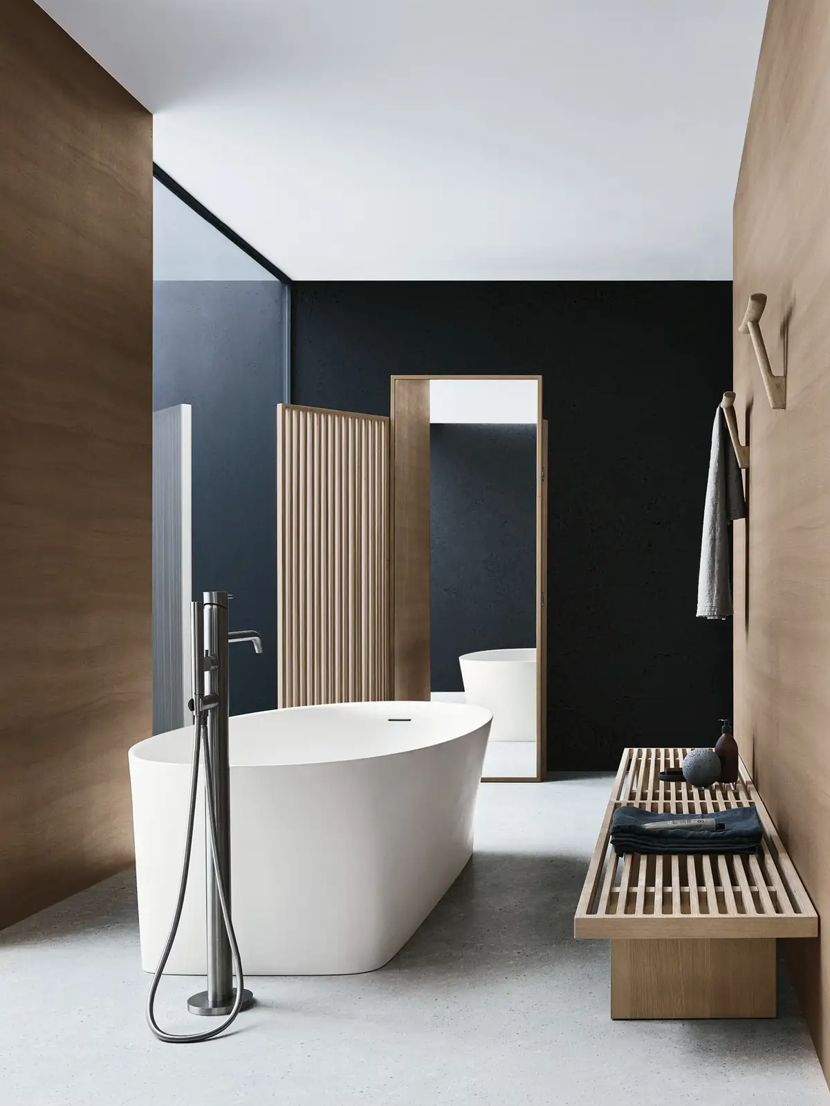 Bañera blanca exenta entre paneles de madera, con un paravento de listones y espejo, un banco de listones y percheros de madera en la pared.