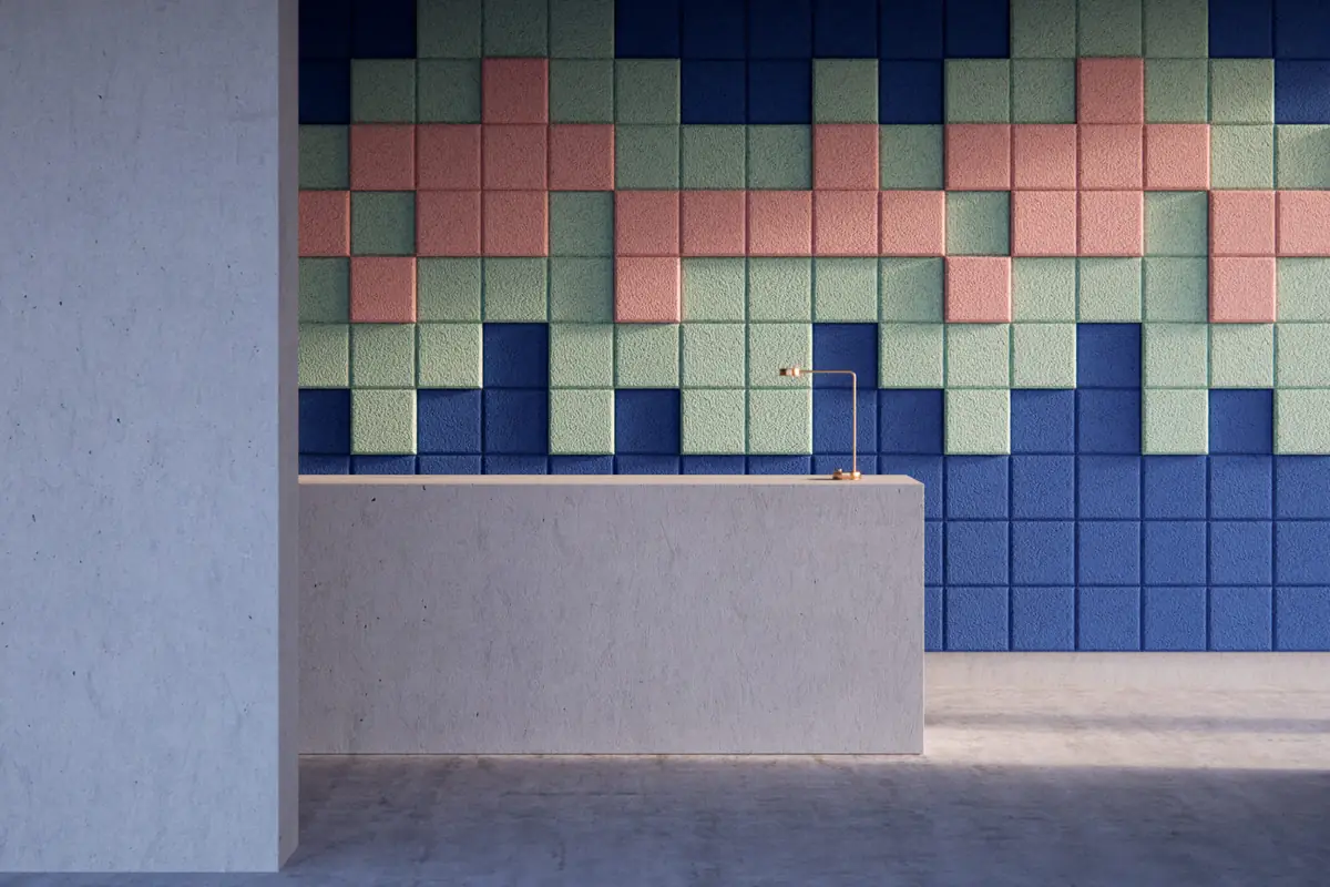 Pared de recepción revestida con baldosas acústicas cuadradas de distintos grosores en azul, verde y rosa tras un mostrador de hormigón