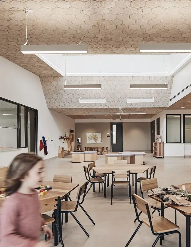 Niña corriendo en una sala con mesas de madera bajo un techo de baldosas hexagonales y un lucernario