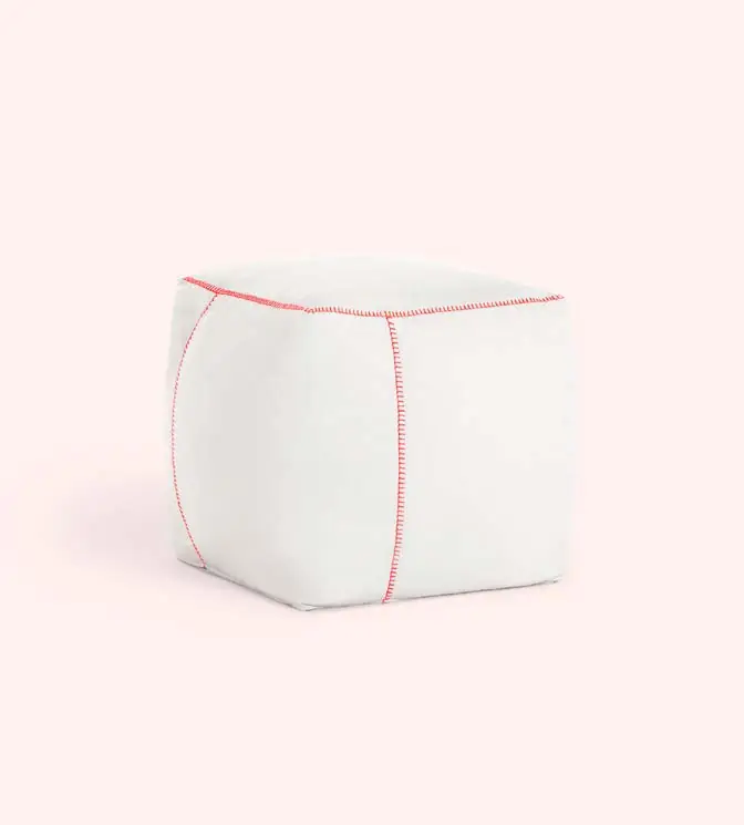 Puf blanco de forma cúbica con costuras rojas en contraste sobre fondo rosa claro