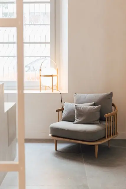 Butaca de madera con cojines de lino gris junto a una ventana y una lámpara de sobremesa