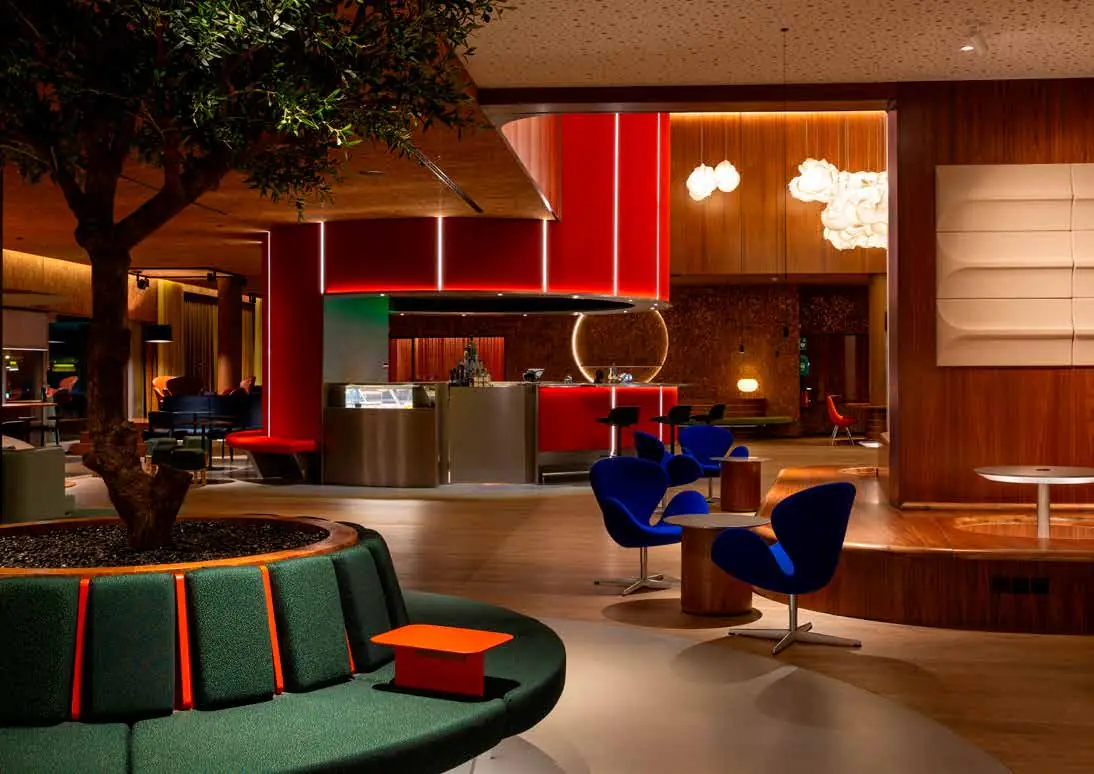 Lobby con un árbol en un banco circular verde, sillones azules y un volumen rojo al fondo