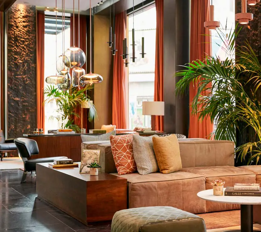 Lobby con sofá de piel, cojines en tonos tierra, cortinas rojizas y plantas