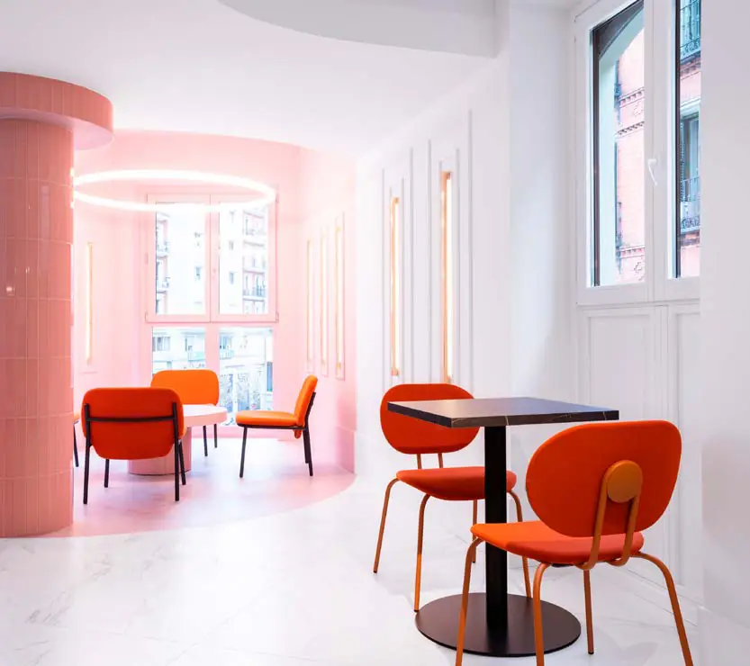 Zona de desayunos con paredes blancas, columna rosa y sillas naranjas