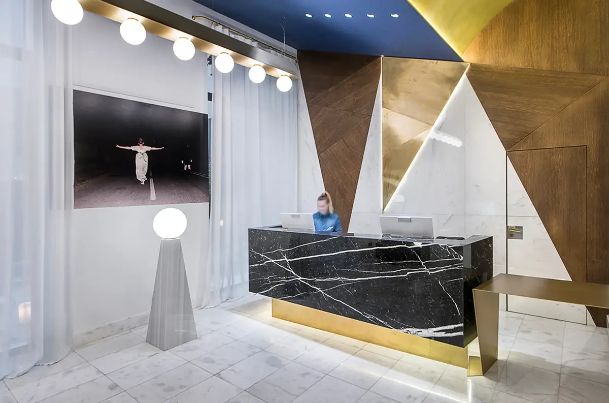 Recepción de hotel con mostrador de mármol negro veteado, paneles triangulares dorados y lámparas de globo
