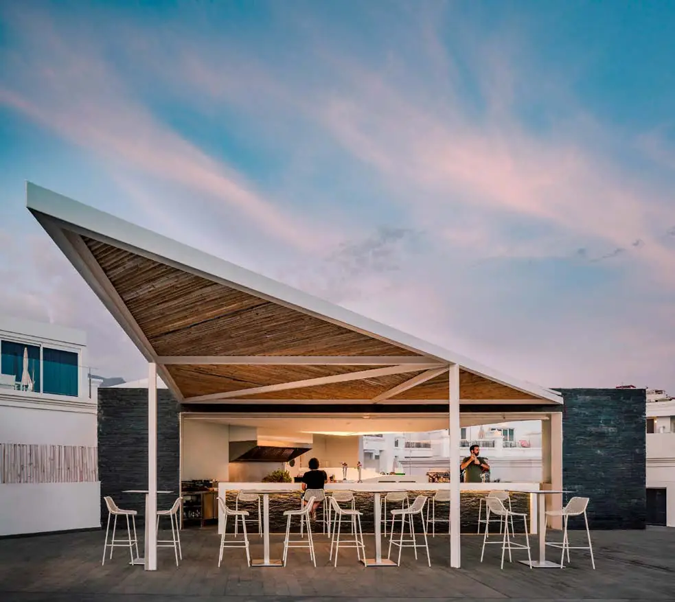 Terraza de restaurante bajo una cubierta inclinada de madera, con mesas altas y fachada blanca