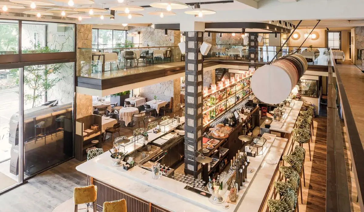 Restaurante de doble altura con barra central, cocina a la vista y grandes ventanales hacia los árboles