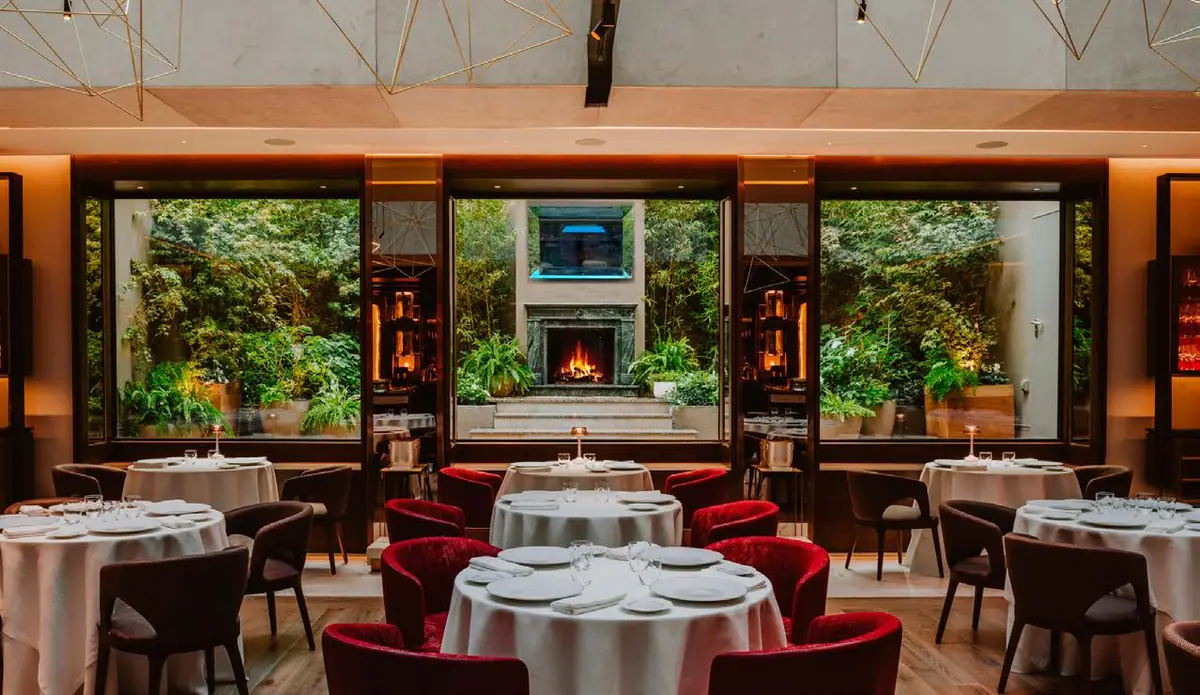 Comedor de restaurante con mesas vestidas de blanco, sillas rojas y ventanales hacia un jardín vertical