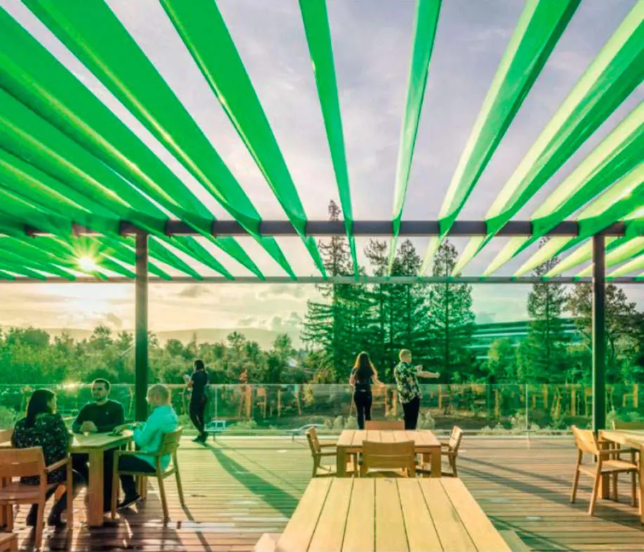 Terraza con mesas de madera bajo una pérgola de lamas verdes translúcidas, con árboles al fondo