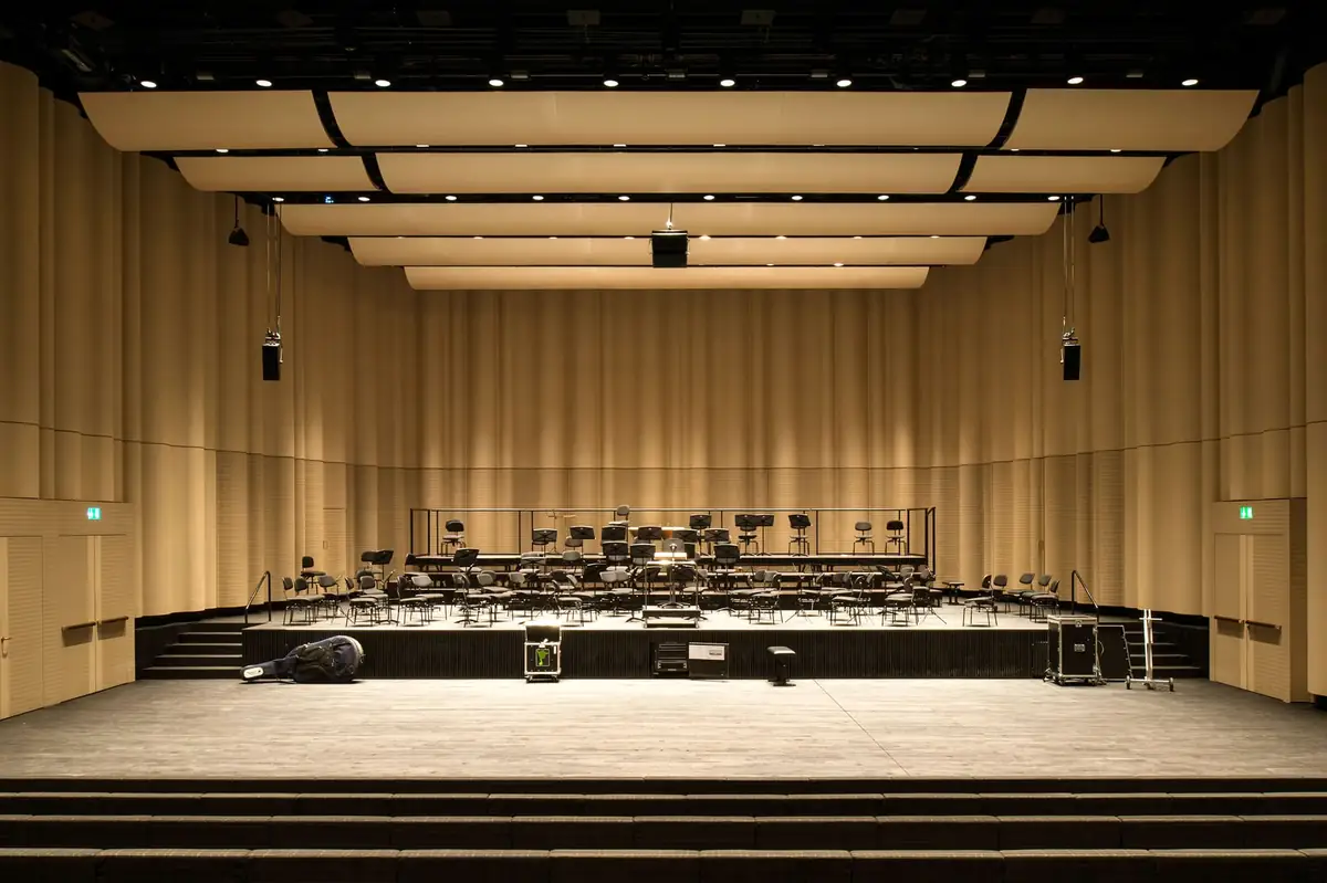 Sala de conciertos con paredes de paneles de madera ondulados en tono claro, escenario con atriles y sillas, y bandejas acústicas suspendidas del techo