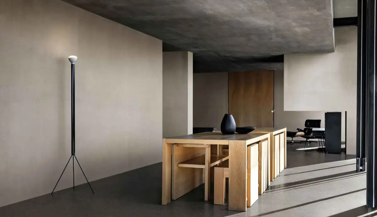 Interior sobrio con paredes y suelo continuos en tonos grises, mesa de madera y lámpara de pie negra.