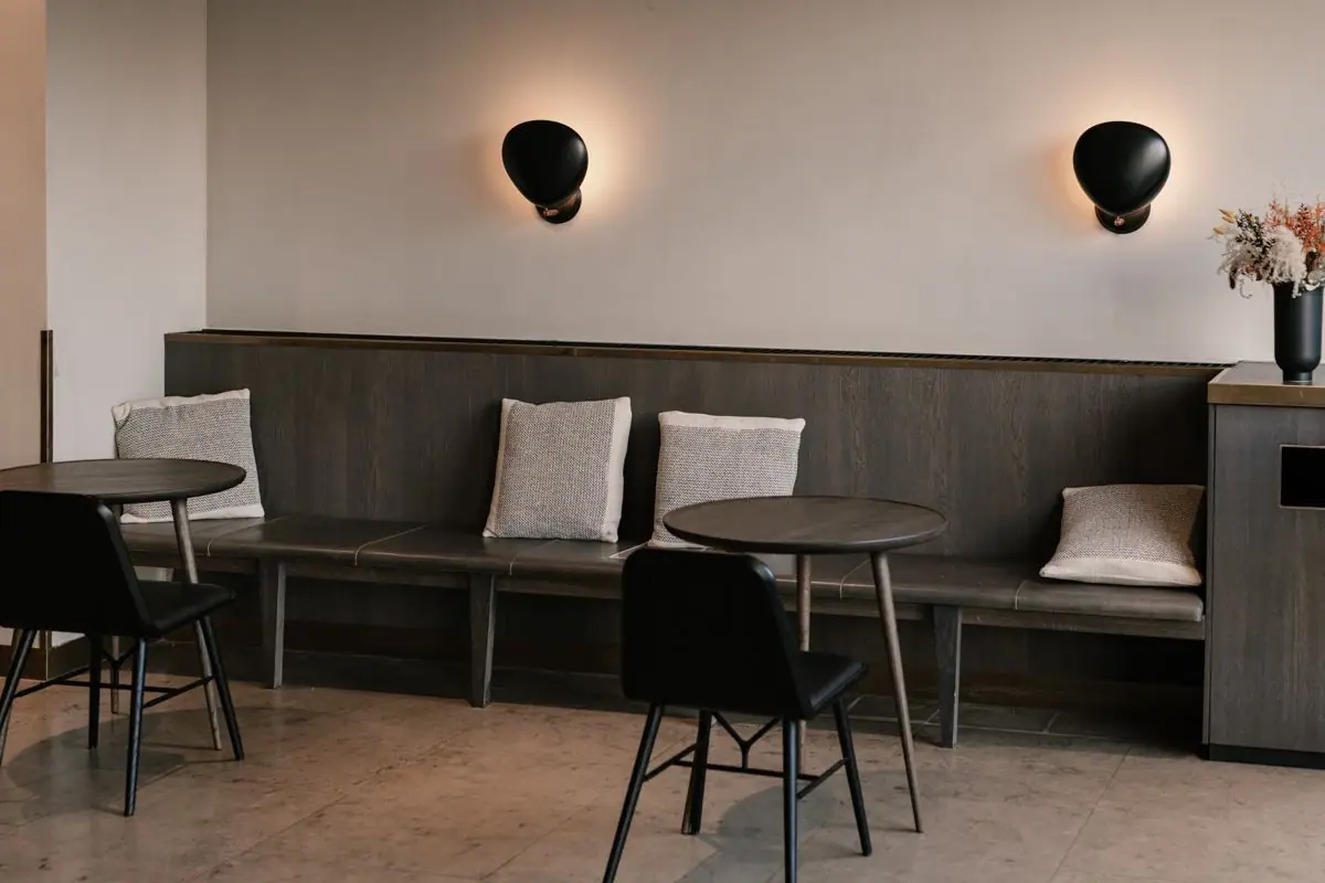 Banco corrido de madera oscura con cojines y dos apliques negros sobre una pared de yeso mineral en tono beige claro