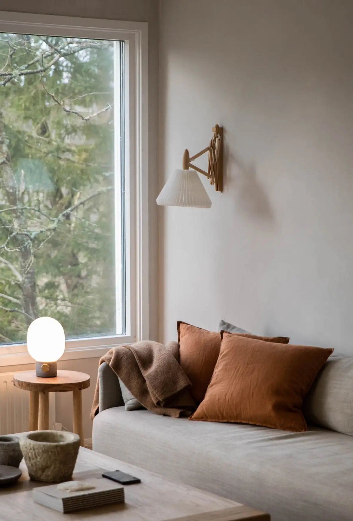 Rincón de salón con ventana, aplique de pared plisado, sofá con cojines terracota y pared de yeso gris claro