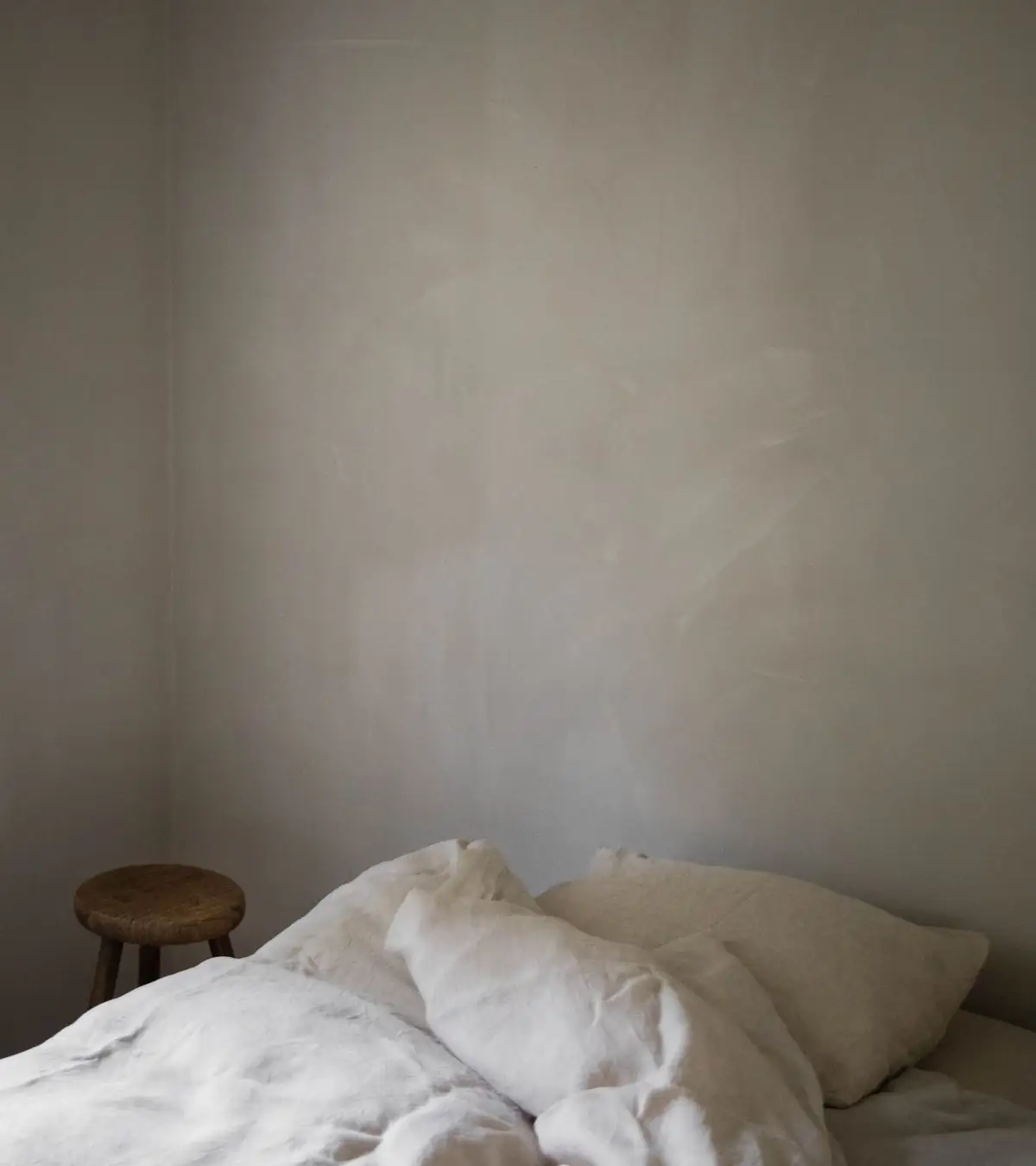 Dormitorio con ropa de cama de lino blanco y taburete de madera ante una pared de yeso con marcas de llana