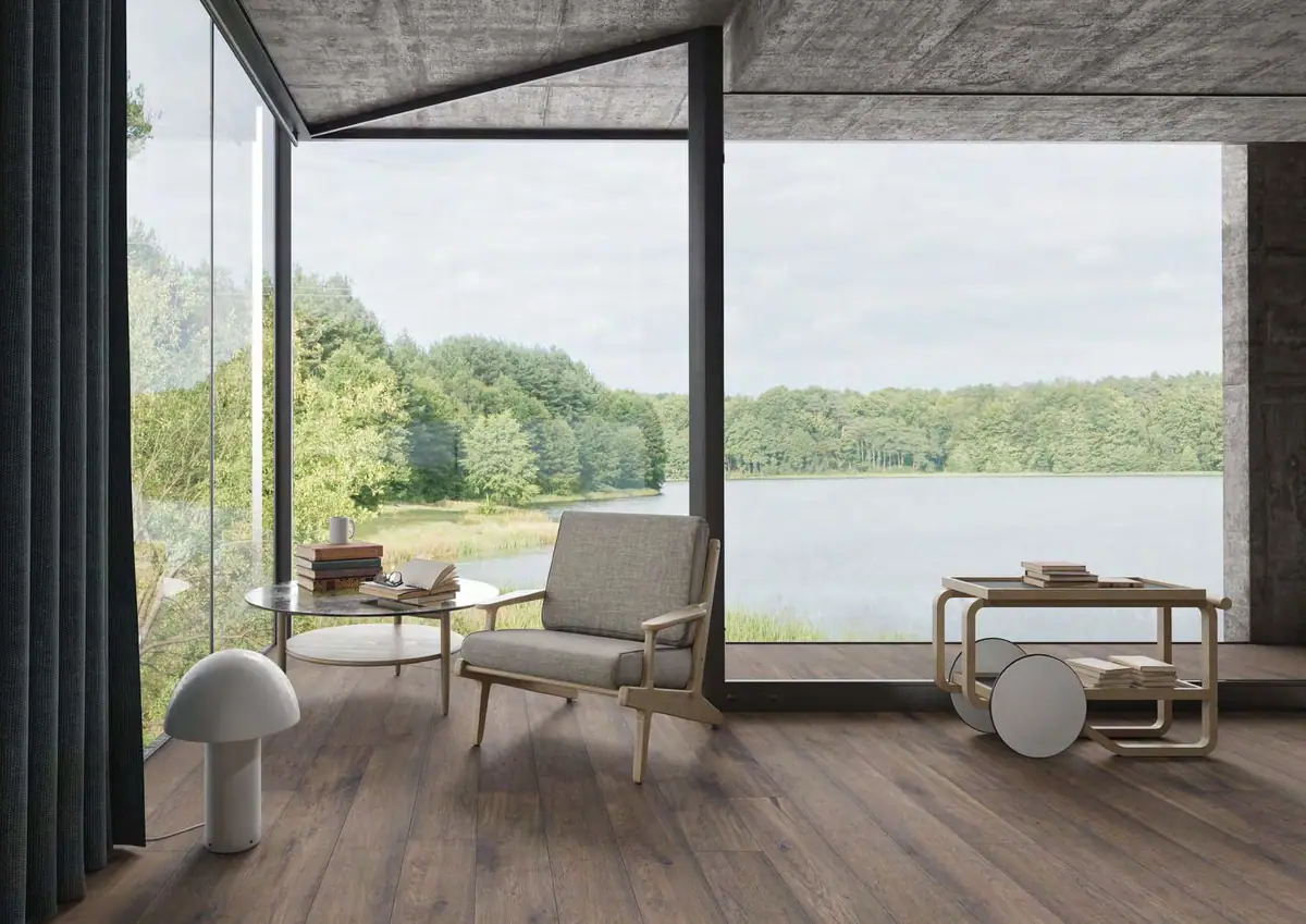 Salón con grandes ventanales sobre un lago, butaca de madera y pavimento de lamas con aspecto de madera oscura