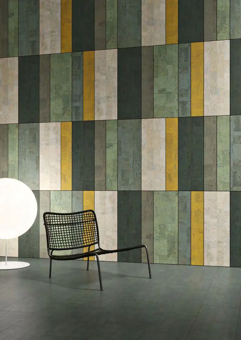 Pared de piezas verticales de corcho en tonos verdes, blancos y amarillo, con una silla de rejilla negra y una lámpara de globo