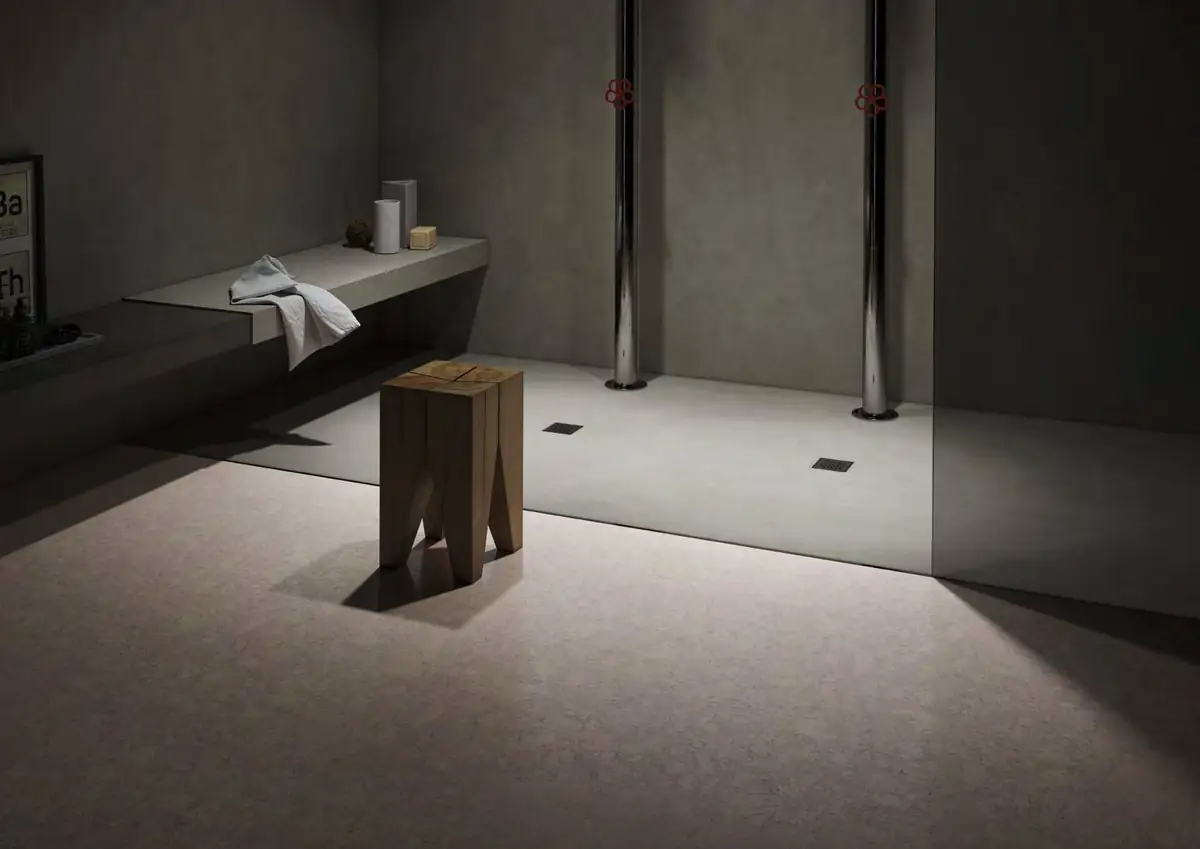 Baño en penumbra con ducha abierta, banco corrido, taburete de madera y pavimento claro de textura moteada