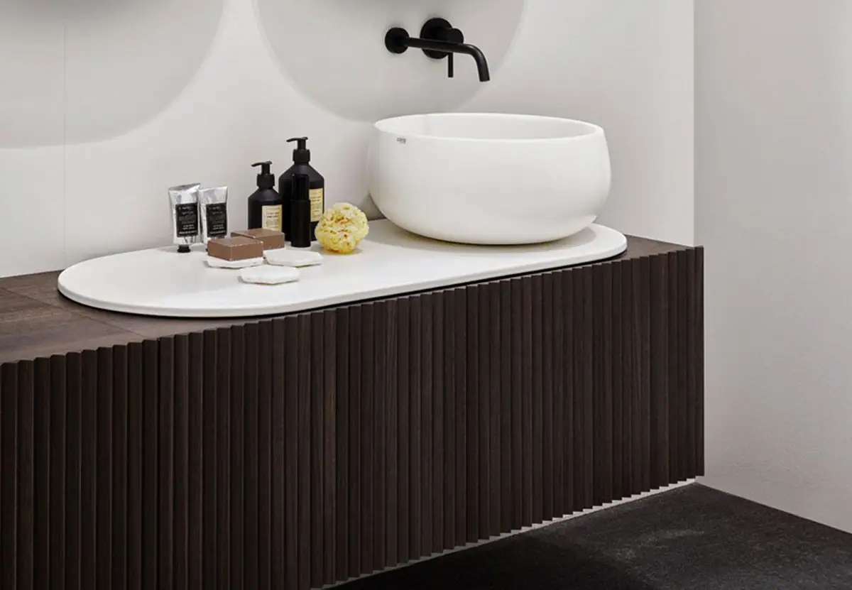 Lavabo de cerámica blanca sobre un top ovalado, encima de un mueble suspendido con frente de lamas de madera oscura y grifo mural negro