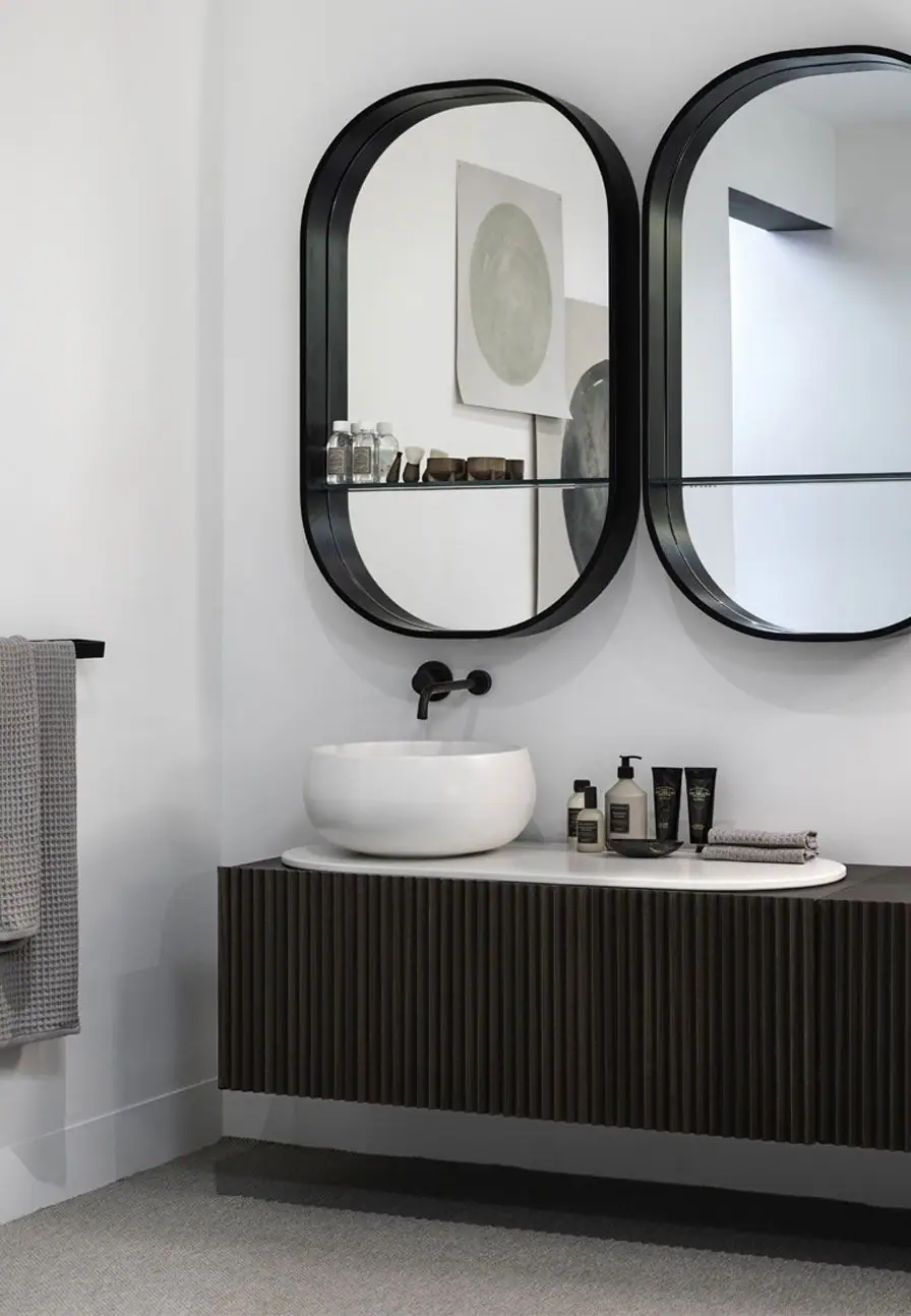 Lavabo blanco sobre top ovalado y mueble suspendido de lamas de madera oscura, bajo dos espejos ovalados de marco negro