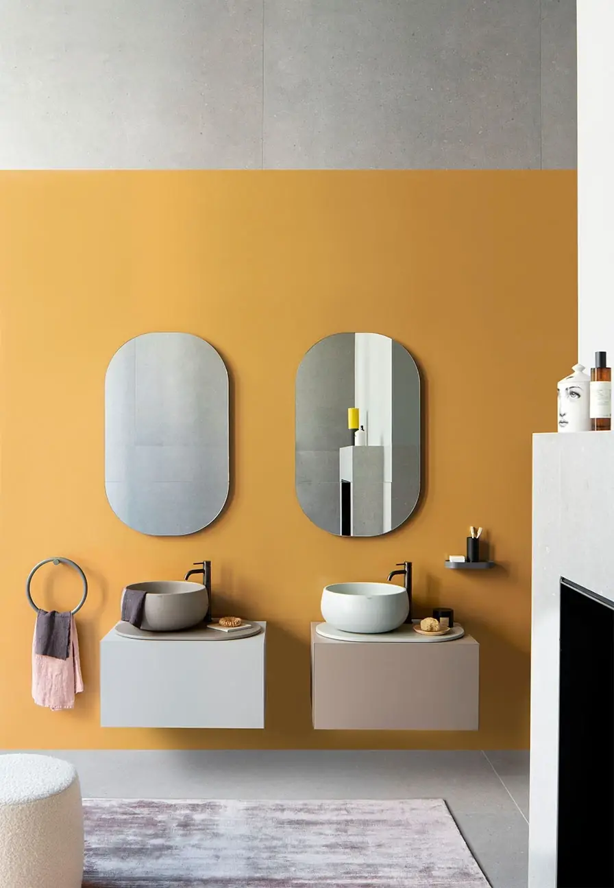 Dos lavabos Delfo 76 con muebles lacados claros y lavabos gris y blanco ante una pared amarilla con espejos ovalados