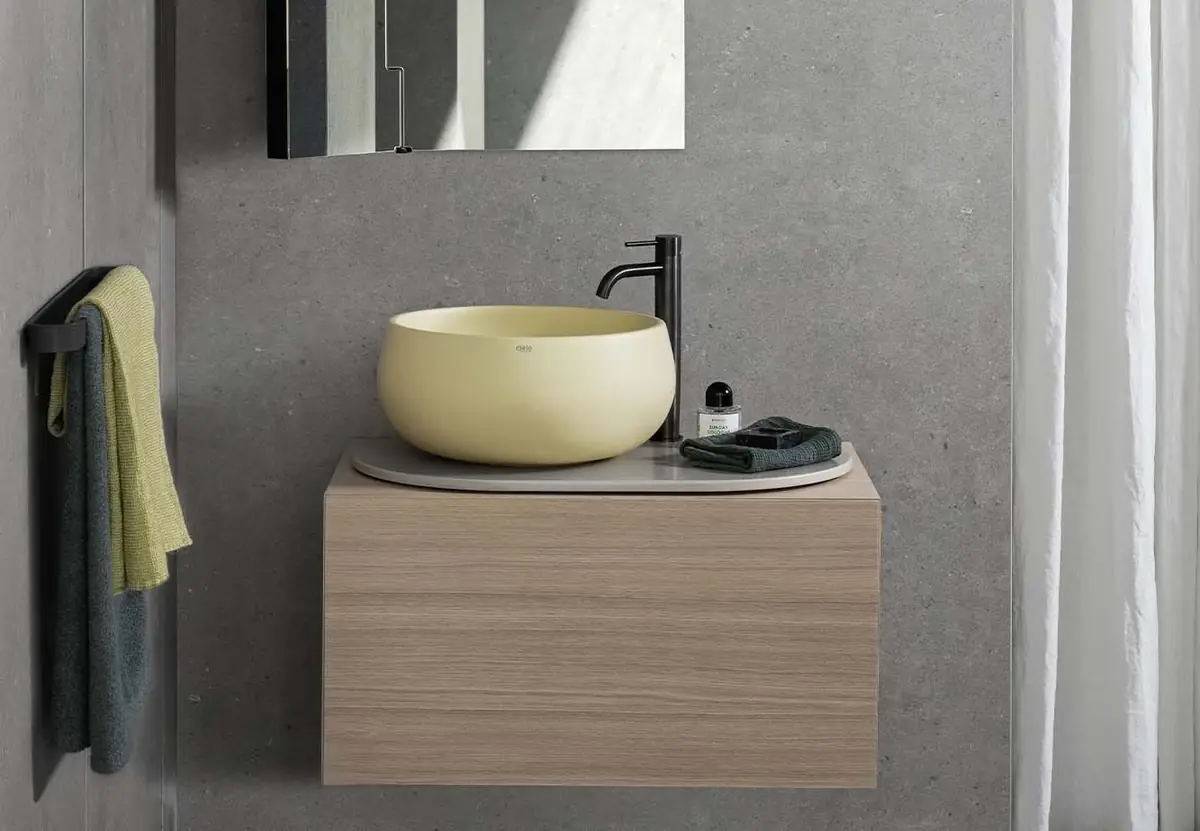 Lavabo de apoyo amarillo pálido sobre un top ovalado gris y un pequeño mueble suspendido de madera clara