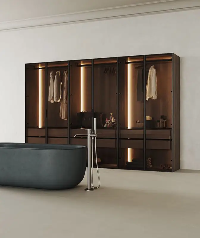 Vestidor con puertas de vidrio y perfiles oscuros, iluminación interior y una bañera exenta gris en primer plano