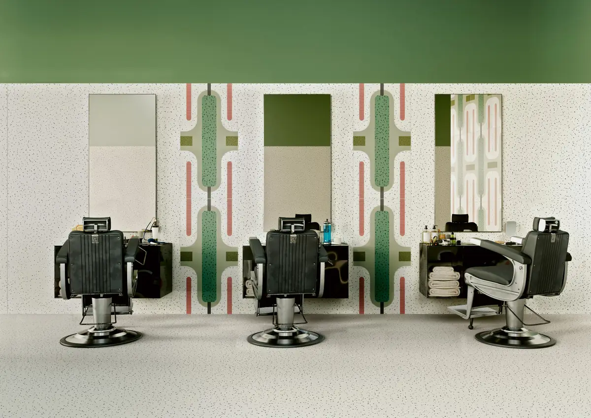 Barbería con tres sillones frente a una pared moteada con bandas decoradas de siluetas verdes y franjas rosas entre los espejos.