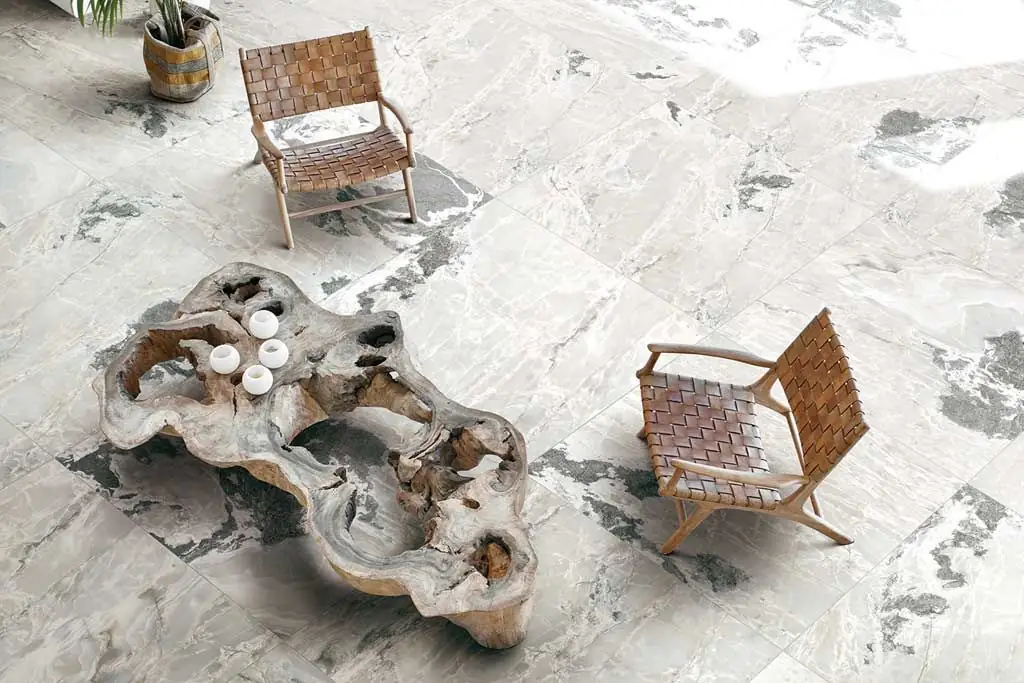 Pavimento de porcelánico de gran formato con efecto de piedra clara y manchas oscuras, con dos butacas de cuero y una mesa de tronco
