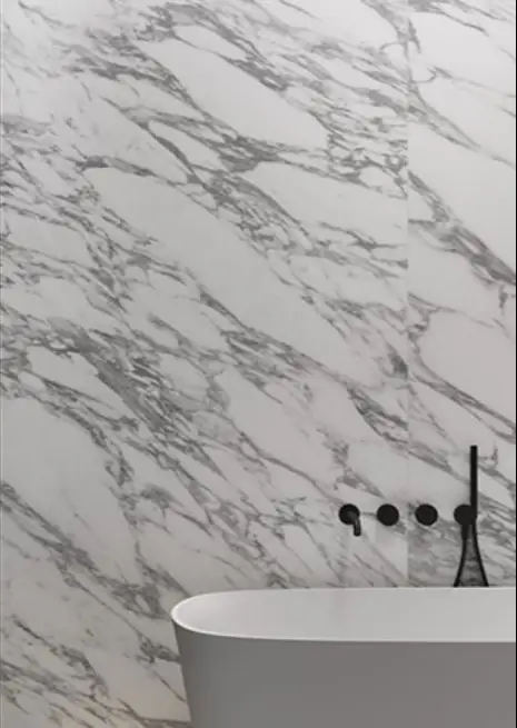 Pared de baño revestida de porcelánico efecto mármol blanco con vetas grises, con grifería negra y bañera blanca