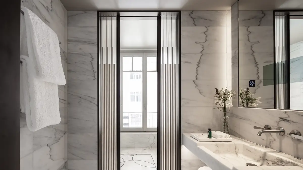 Baño revestido de mármol blanco veteado con lavabo de mármol y grifería de pared en acero, junto a una puerta de vidrio acanalado
