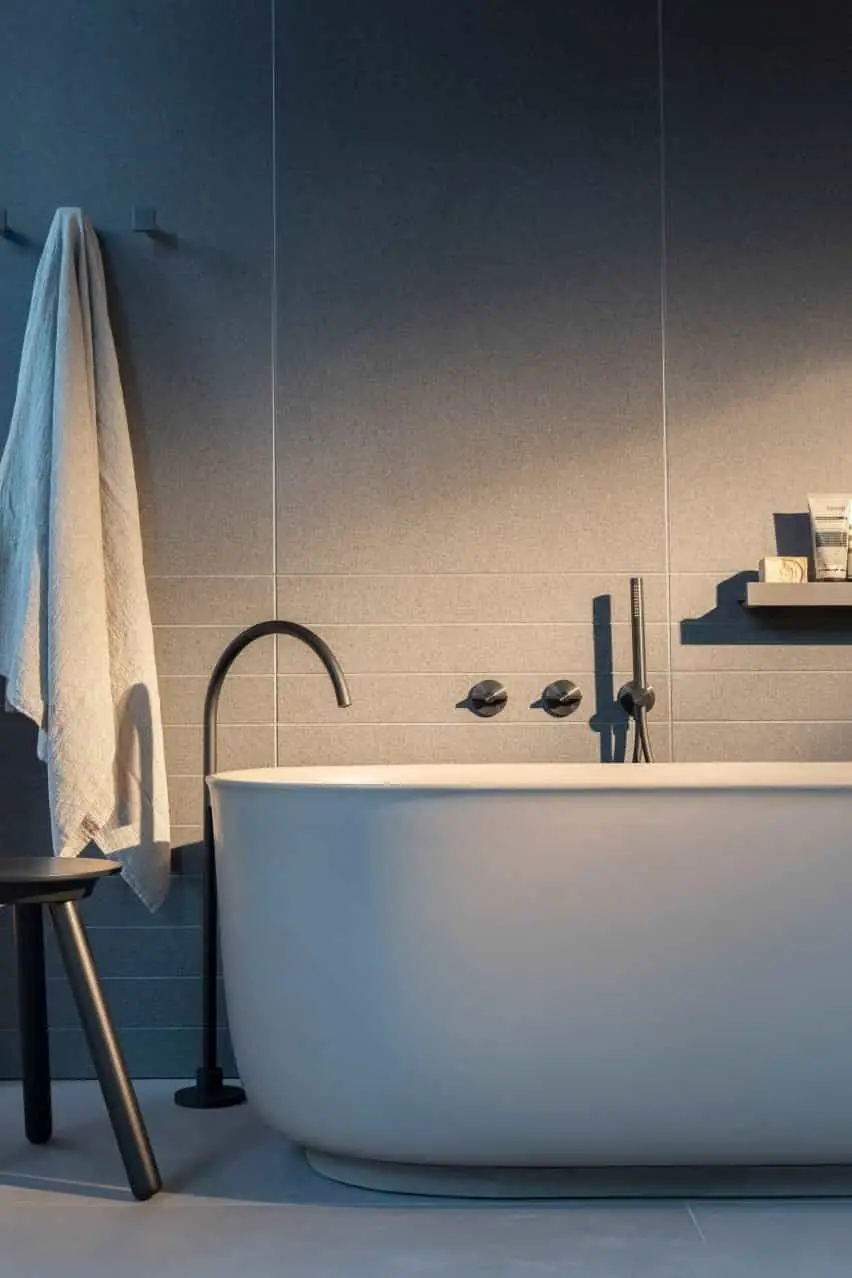Bañera exenta blanca con caño de pie negro y mandos y ducha de mano en la pared, junto a una toalla y un taburete