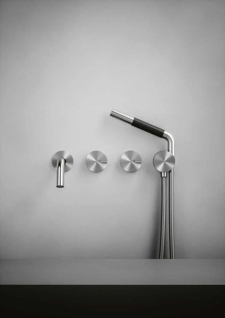 Caño de pared, dos mandos circulares y ducha de mano de acero inoxidable sobre una pared gris