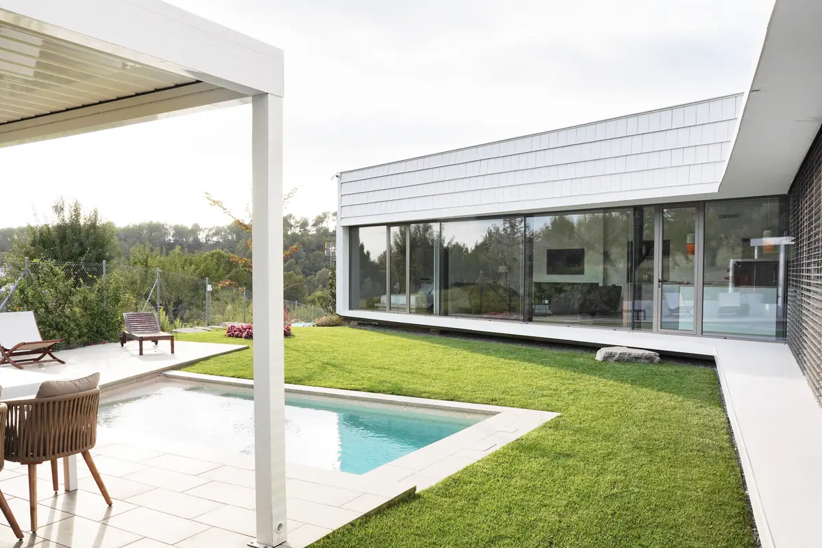Jardín y piscina de la casa Om, con la fachada acristalada de la vivienda y el revestimiento blanco de tablillas en la parte superior