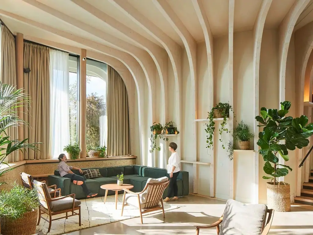 Sala interior de doble altura con arcos de madera clara, grandes ventanales, sofá verde y plantas.