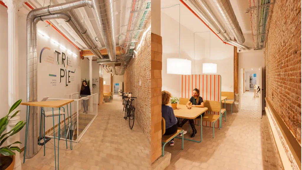Planta baja de Triple: pasillo con muro de ladrillo visto, rótulo del coworking, bicicletas y una zona de mesas de madera con bancos