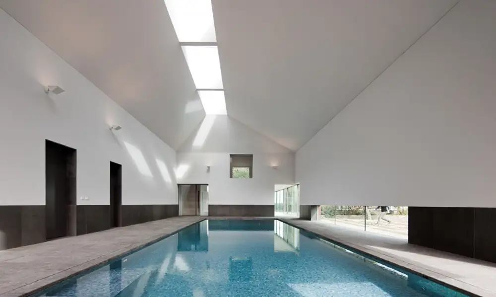 Piscina cubierta con techo inclinado blanco, lucernario longitudinal y zócalo oscuro.