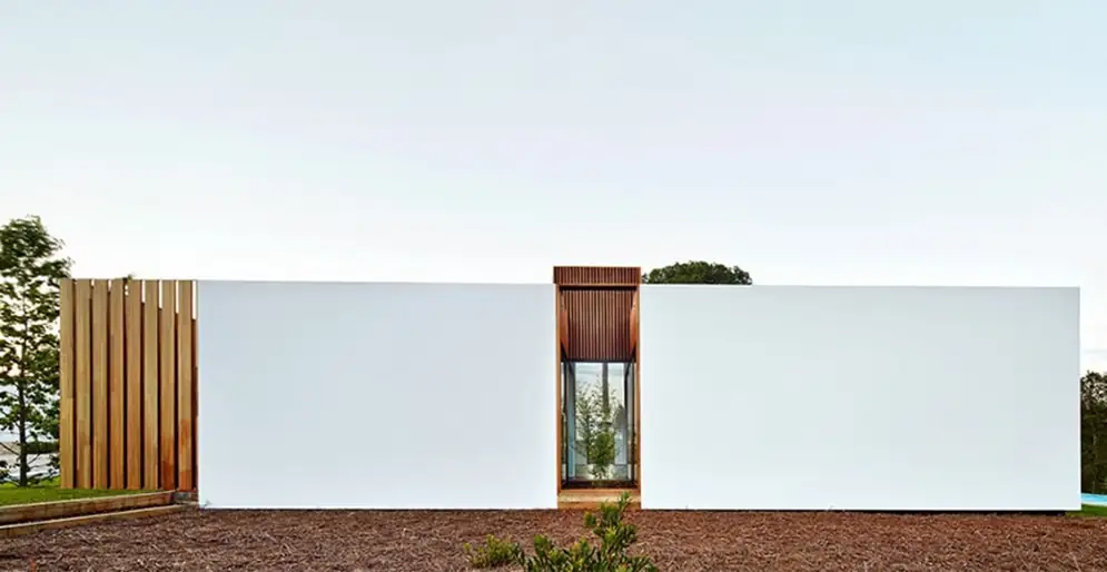 Vivienda de una planta con fachada blanca continua, una entrada vertical de madera y celosía de lamas de madera.