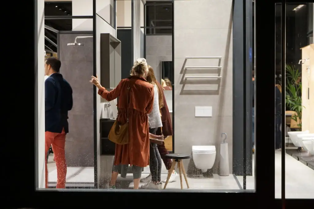 Invitados observando un microambiente de baño con inodoro suspendido y toallero, visto a través del cristal