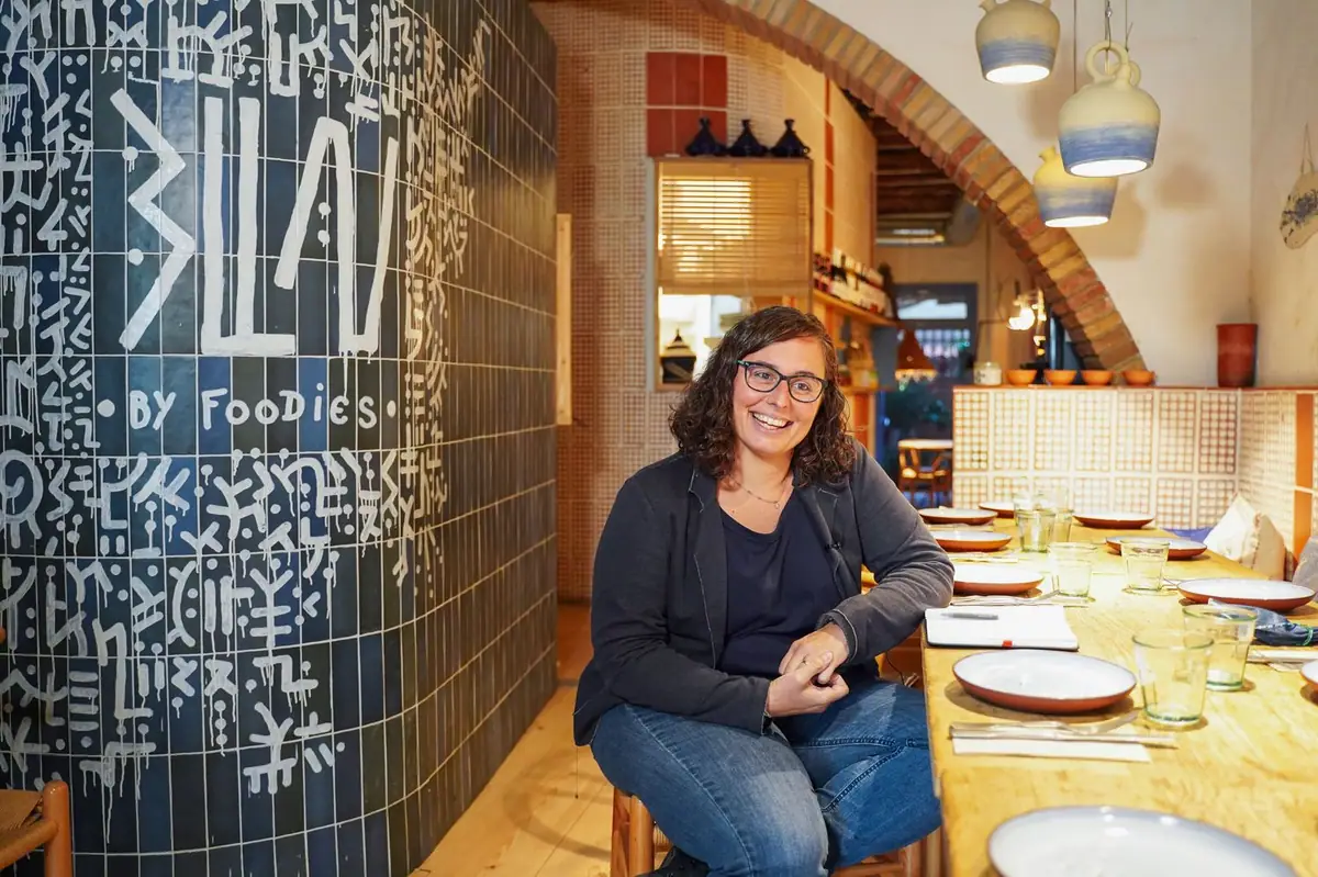 Una persona sentada junto a una pared de azulejo azul con el rótulo Blau by Foodies y una mesa corrida de madera