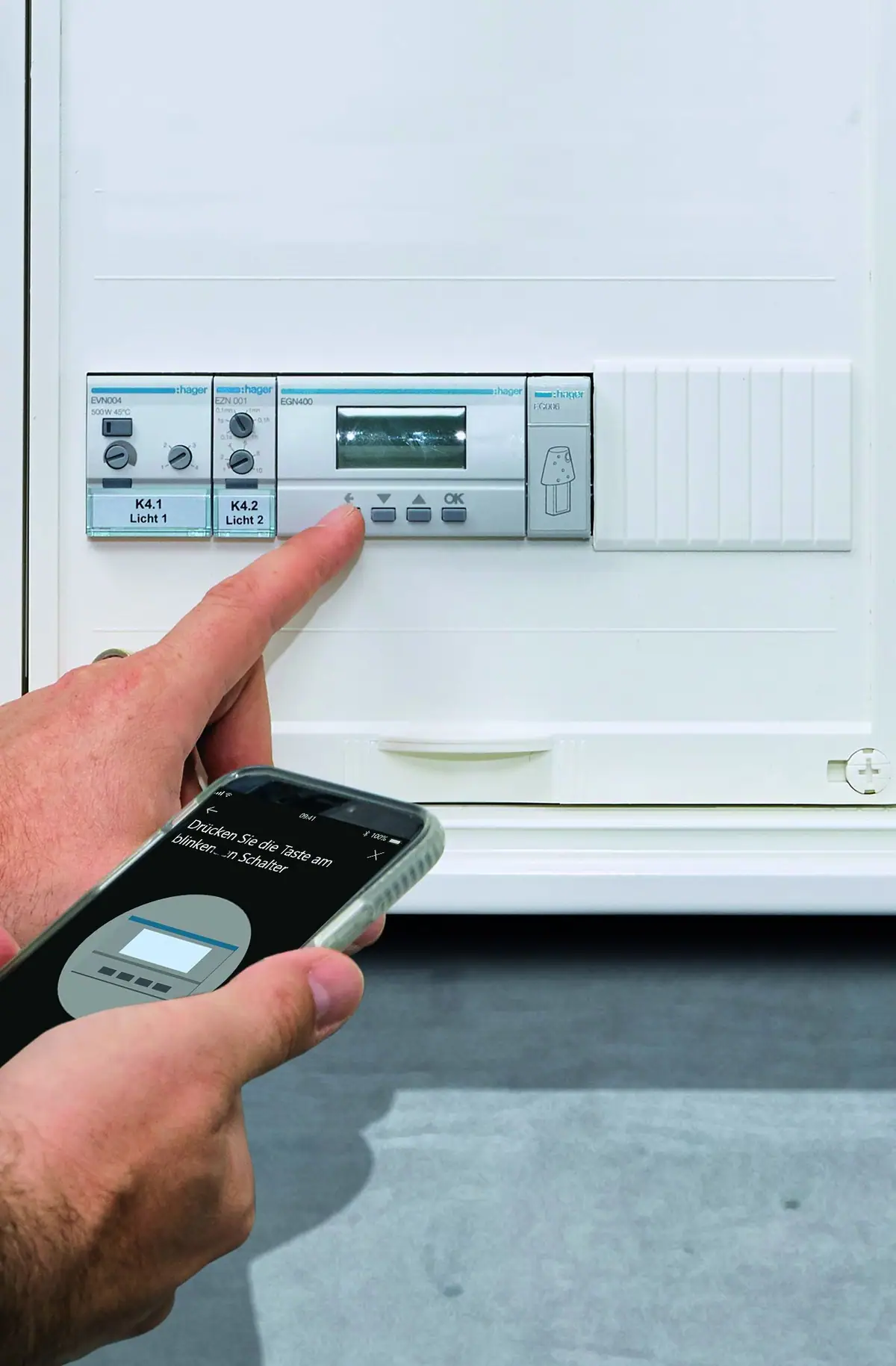 Mano que pulsa un programador horario digital Hager EGN400 en un cuadro eléctrico mientras otra sostiene un móvil con la aplicación de configuración