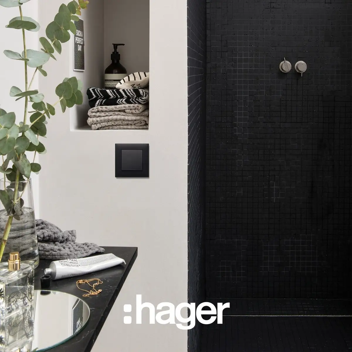 Interruptor negro en la pared de un baño, junto a una ducha revestida de mosaico negro y un estante con toallas