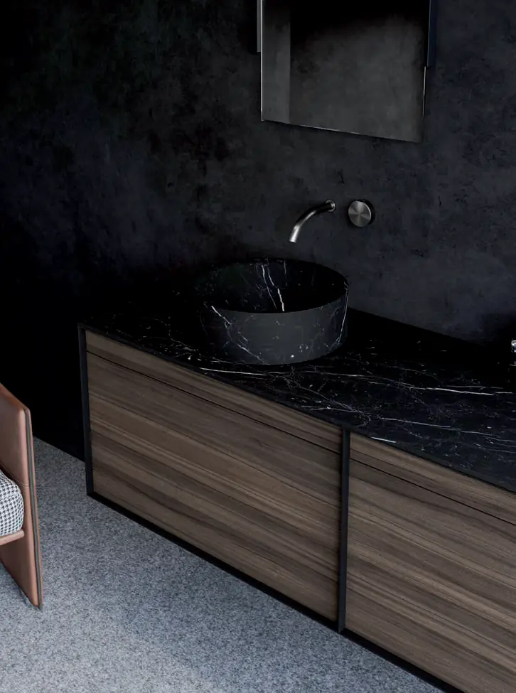 Mueble con frentes de madera natural, encimera de mármol negro veteado y lavabo de apoyo a juego, en un baño de paredes oscuras.