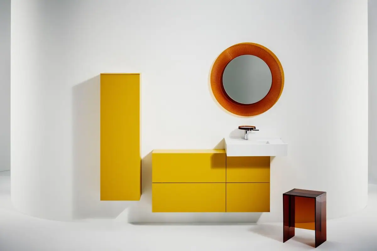 Columna y muebles bajo lavabo amarillos con lavabo blanco, espejo redondo naranja y taburete transparente ámbar