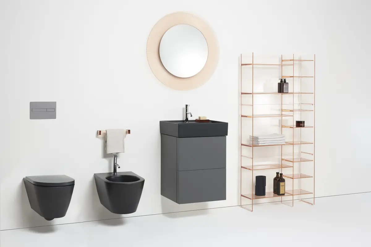 Inodoro y bidé suspendidos oscuros, mueble de lavabo gris, espejo redondo rosado y estantería transparente cobre